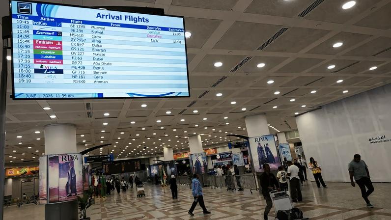 Passengers walk past a screen displaying flight arrival information at Kuwait International Airport.