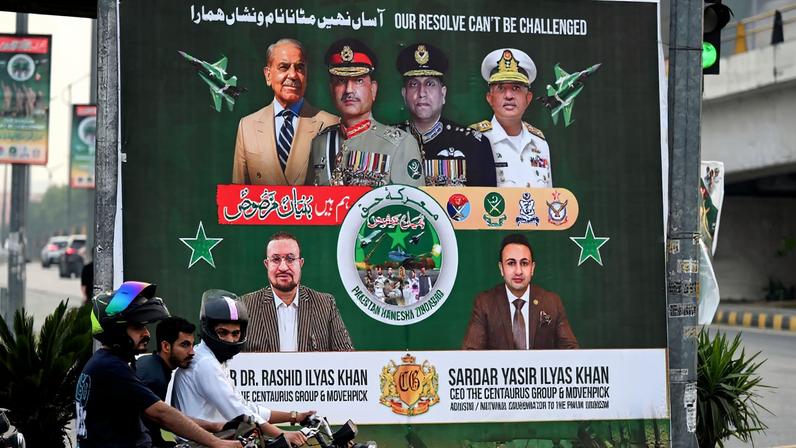 Commuters wait at a traffic signal next to a billboard featuring, from left to right, Pakistani Prime Minister Shehbaz Sharif, Army chief Asim Munir, Air Chief Marshal Zaheer Ahmad Babar, and Navy chief Adm. Naveed Ashraf, displayed to mark a year since the India-Pakistan conflict, along a street in Islamabad.