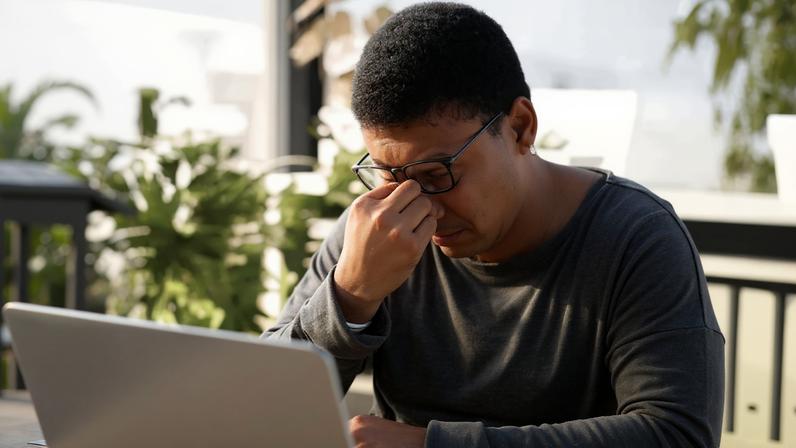 Imagem de um rapaz, diante de um notebook, com semblante cansado e coçando os olhos.