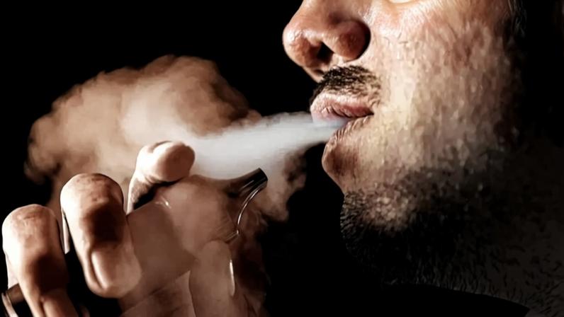 Close-up de uma pessoa exalando fumaça de um vape, com a boca e parte do rosto visíveis. A fumaça branca se espalha no ar escuro, enquanto um dispositivo vape é segurado por uma mão. Há blocos de cores sólidas em amarelo, rosa e azul no fundo.