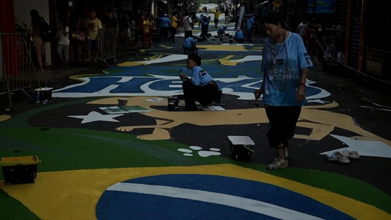 Pessoas pintam a rua com as cores da bandeira do Brasil: verde, amarelo, azul e branco. Uma mulher de camiseta azul e calça escura caminha com um pincel, enquanto outra, agachada, pinta uma estrela branca. Ao fundo, mais pessoas participam da pintura e outras observam. A rua está decorada com desenhos abstratos e elementos da bandeira brasileira.