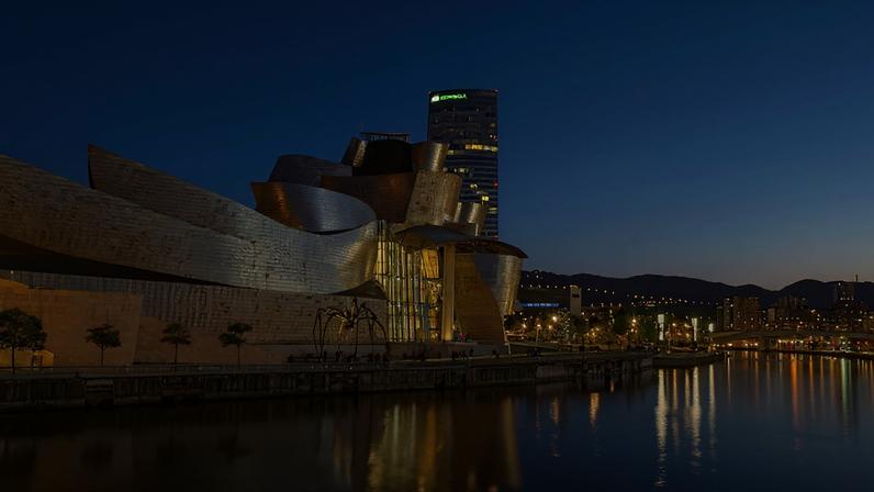 Imagem do Museu Guggenheim Bilbao, com sua arquitetura metálica curvilínea, reflete a luz do entardecer sobre o rio. Ao fundo, um arranha-céu e a cidade iluminada se estendem até as montanhas. Uma escultura de aranha gigante está em frente ao museu.