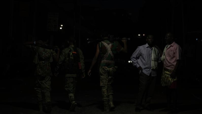 Three soldiers are shown on a street from behind, while pedestrians stand near them.