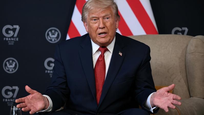 U.S. President Donald Trump sits on a chair while addressing the media. His hands are gesturing on either side of him. A U.S. flag is seen in the background.