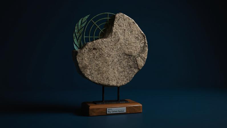 A studio shot of an artifact displayed on a small wooden base against a solid blue background. The artifact features a large, rough-textured stone disk with an uneven edge. Embedded or emerging from the upper-left edge of the stone is a green, weathered metal emblem resembling the United Nations logo, including an olive branch and a partial grid pattern. The stone structure is mounted on two short black metal pillars attached to a polished wooden stand, which has a small white label on the front that reads "EXHIBIT 1: The United Nations".