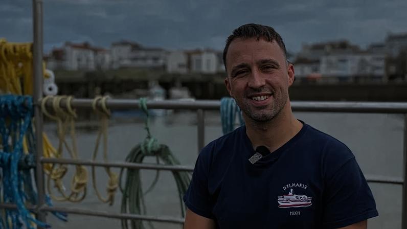 BBC A man in a blue t-shirt is standing in front of railings, with a harbour in the background. He has short brown hair and stubble. His T-shirt features a logo of a fishing boat and the name Dylharis. Ropes in blue, yellow and green are attached to the railings.