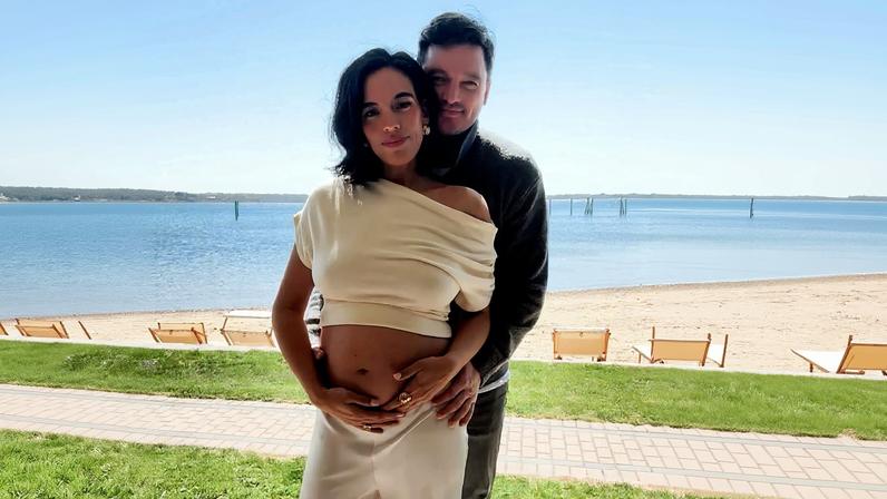 Danielle Olivera and Eoin Heavey posing with her baby bump, with a beach and water in the background.