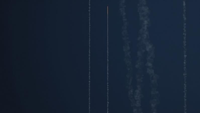 A rocket is seen from a great distance, flying through a deep blue sky during military drills. Several parallel plumes of smoke are seen alongside the rocket's path.