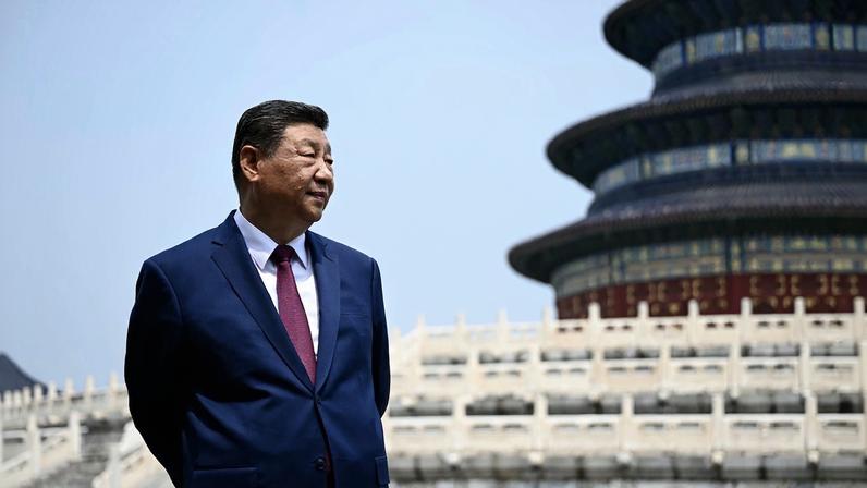 Chinese President Xi Jinping, wearing a dark blue suit with a red tie, stands in front of the Temple of Heaven in Beijing, looking onward.