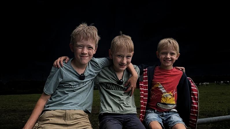 BBC Three children sit on top of a gate in a field with cows in. They are smiling at the camera. On the left is a blond haired boy wearing a blue t-shirt and beige shorts. In the middle another blond boy wears a green t-shirt and navy shorts and on his right is another blond haired younger boy wearing a red t-shirt, denim shorts and a red stripey jacket.