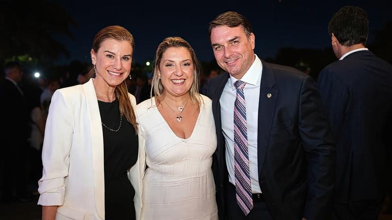 Daniella Marques entre o casal Flávio Bolsonaro e Fernanda durante evento da Caixa, em 2022