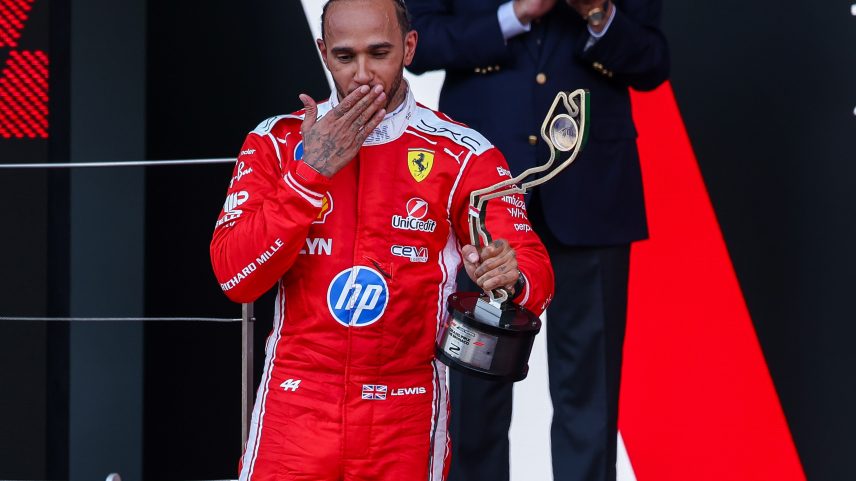 Lewis Hamilton in a red racing suit blows a kiss while holding a trophy at the Monaco Grand Prix.