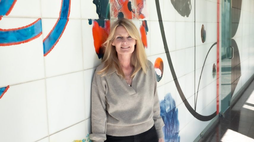 Karen O’Leary stands against a painted tile wall in well-lit hallway