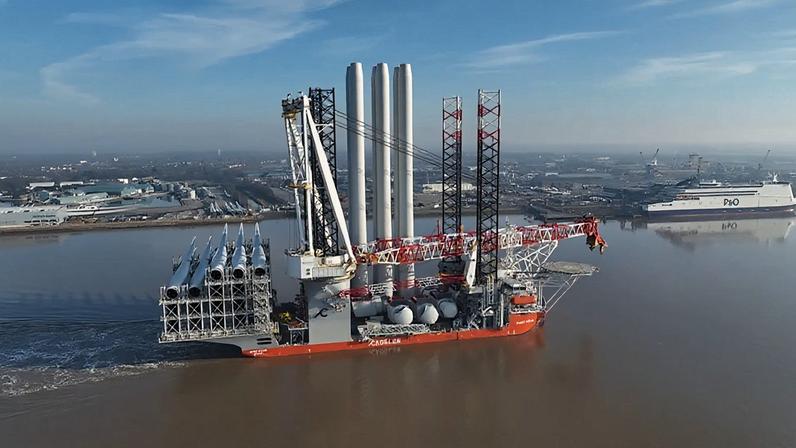 A massive offshore wind installation ship carrying turbine components sails along a river near an industrial port.