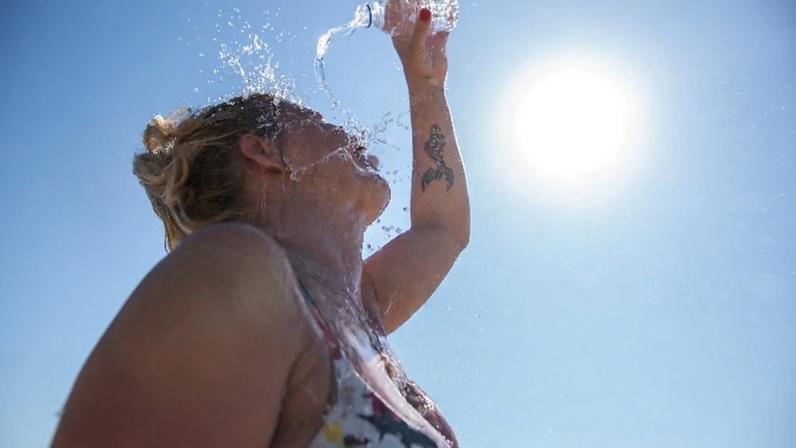 Woman pours a bottle of water over her head as she stands in sun rays.