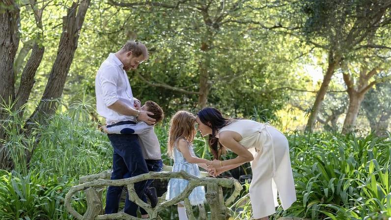 Prince Harry, Meghan Markle, and their two children, Archie and Lilibet, standing on a bridge in a lush garden.
