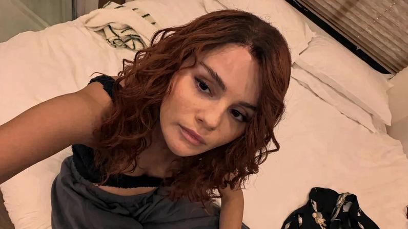 Selena Gomez with curly red hair, looking up at the camera while sitting on a white bed.
