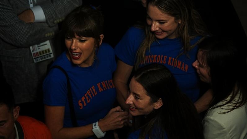 Taylor Swift in a blue "STEVIE KNICKS" t-shirt, laughing with other people, some of whom are wearing "NICKOLE KIDMAN" shirts.