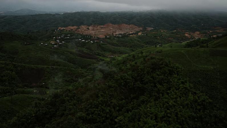 A wide aerial shot shows a mine from a distance, beyond a stretch of rolling green hills. The mine is a pale brown area among the green, stripped of vegetation, made up of large mounds of pale brown dirt and the small valleys between them.