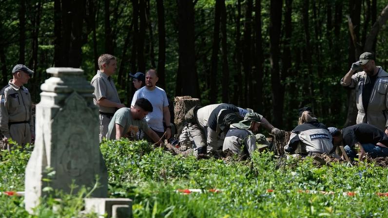 About a dozen people are seen across a space overgrown with short green plants rising to about knee height. A few people lean down to examine the ground, and some wear gloves or coveralls that identify them as archaeologists. In the foreground is a gray obelisk, either a grave marker, another memorial, or part of a building that is no longer extant.