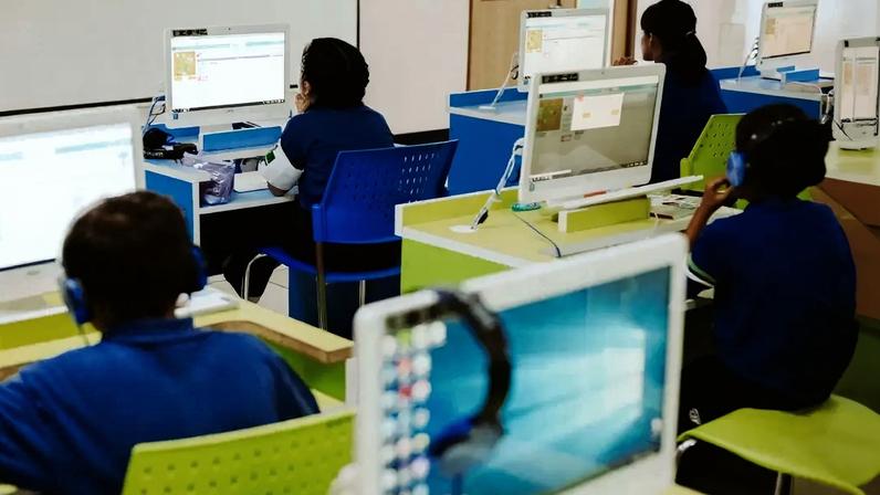 Alunos em uma sala de aula de informática, sentados em mesas coloridas com computadores e fones de ouvido, focados nas telas
